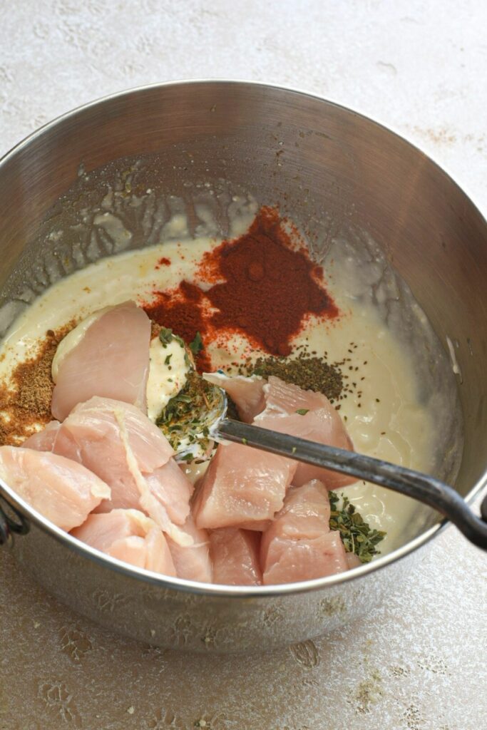 A large metal bowl filled with cubed raw chicken that is about to be mixed with yogurt, celery seed, paprika, italian seasoning and other spices