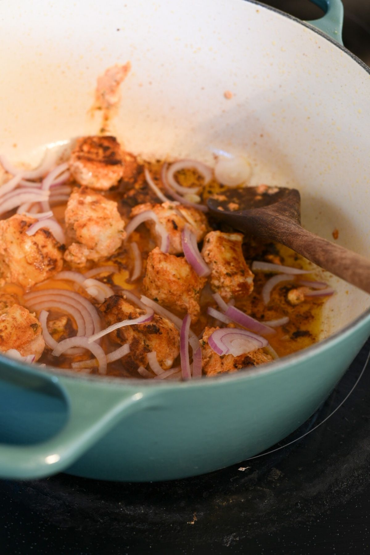 A large blue dutch oven filled with sauteing chicken cubes and red onion. There is a wooden spoon in the pot.