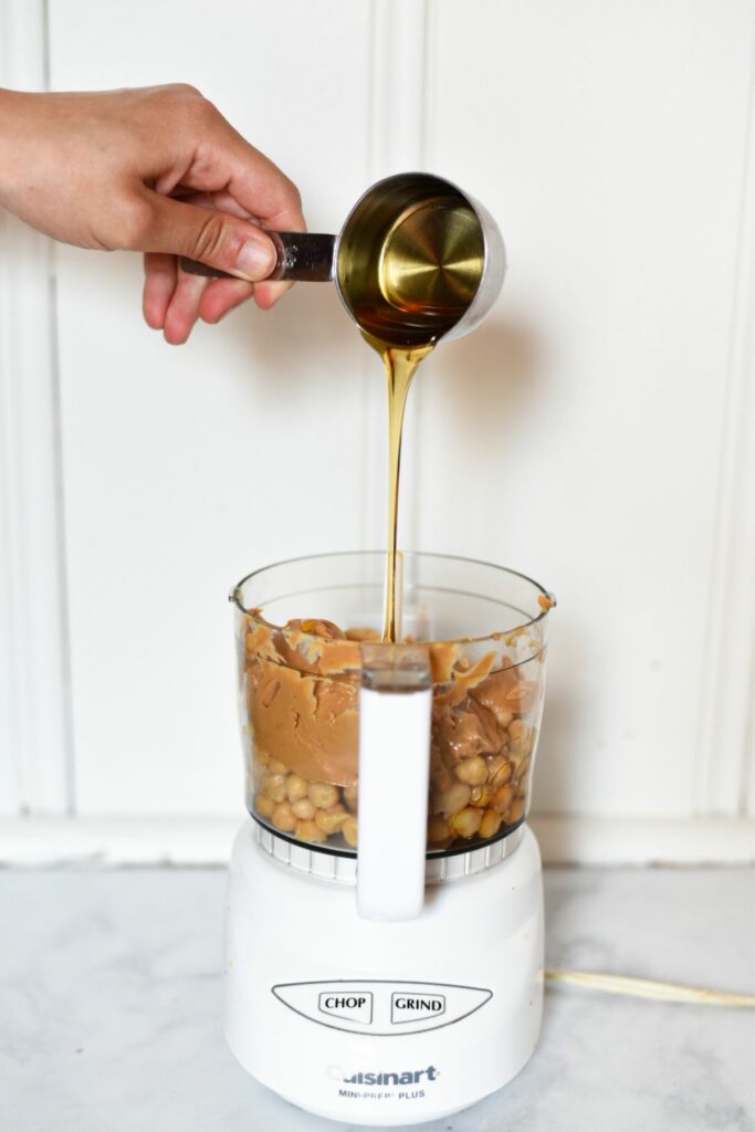 A hand pouring honey into a food processor filled with chickpeas and peanut butter