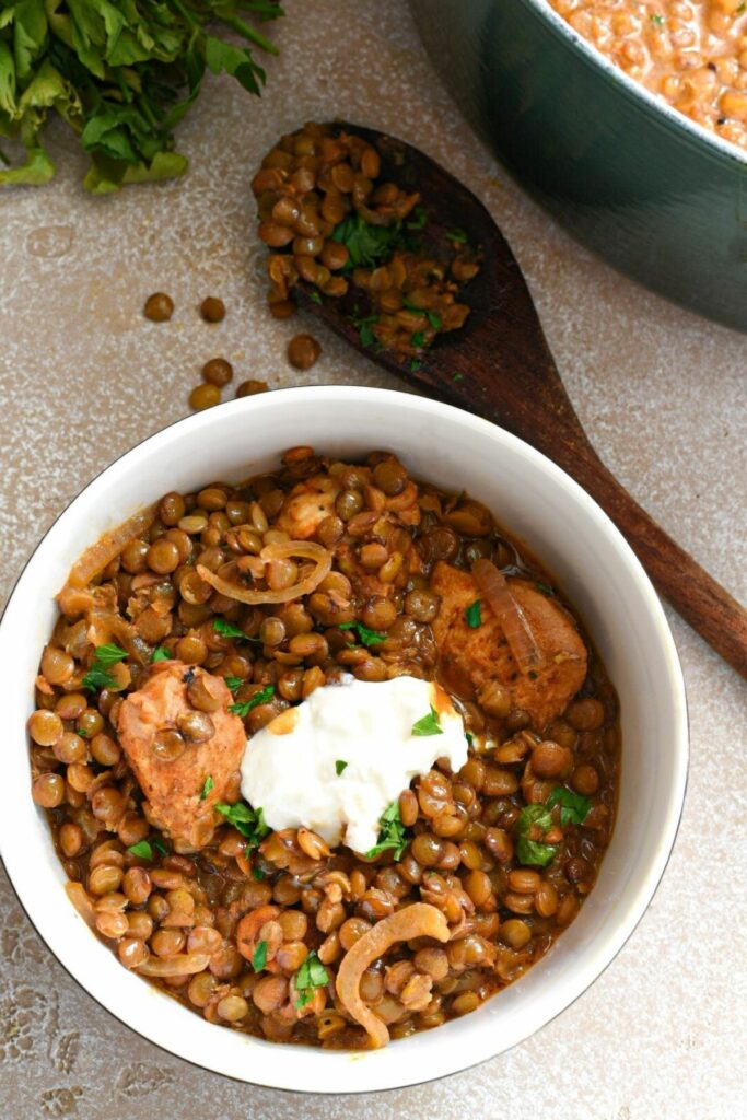 A close up overhead shot of a bowl filled with cooked lentils, with pieces of cubed marinated chicken showing, a dollop of white yogurt, and parsley sprinkled over the top.