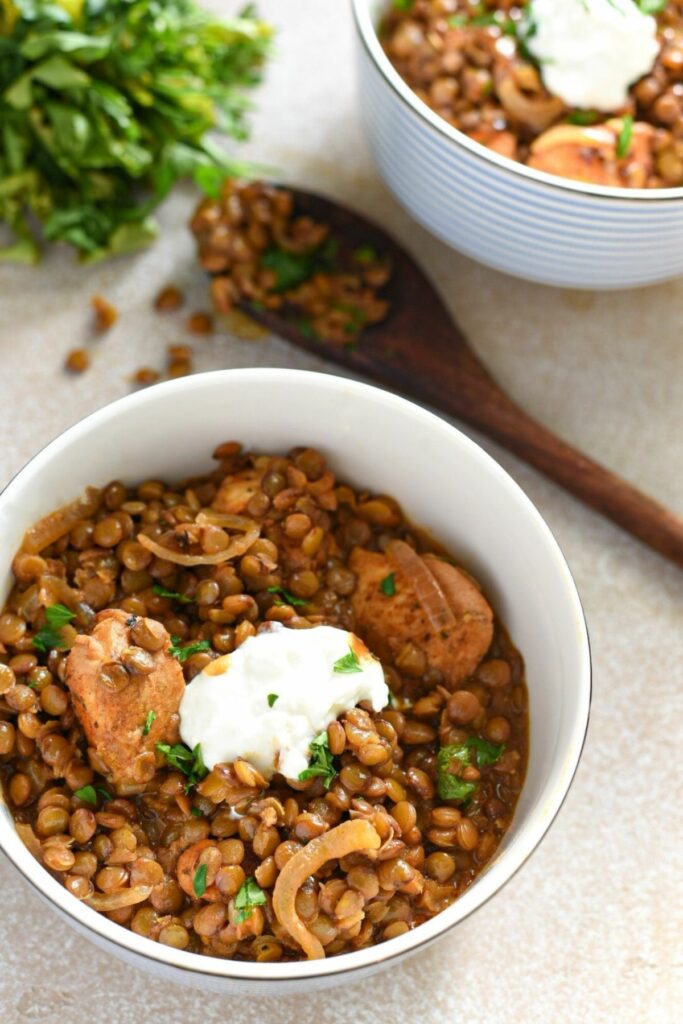 A small bowl filled with brown lentil stew, with pieces of cooked marinated chicken, yogurt and parsley on top