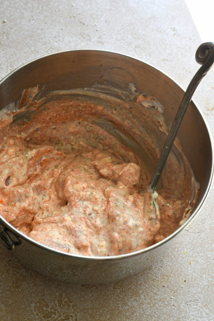 A metal bowl filled with cubed raw chicken, marinating in a red yogurt sauce with paprika and other spices