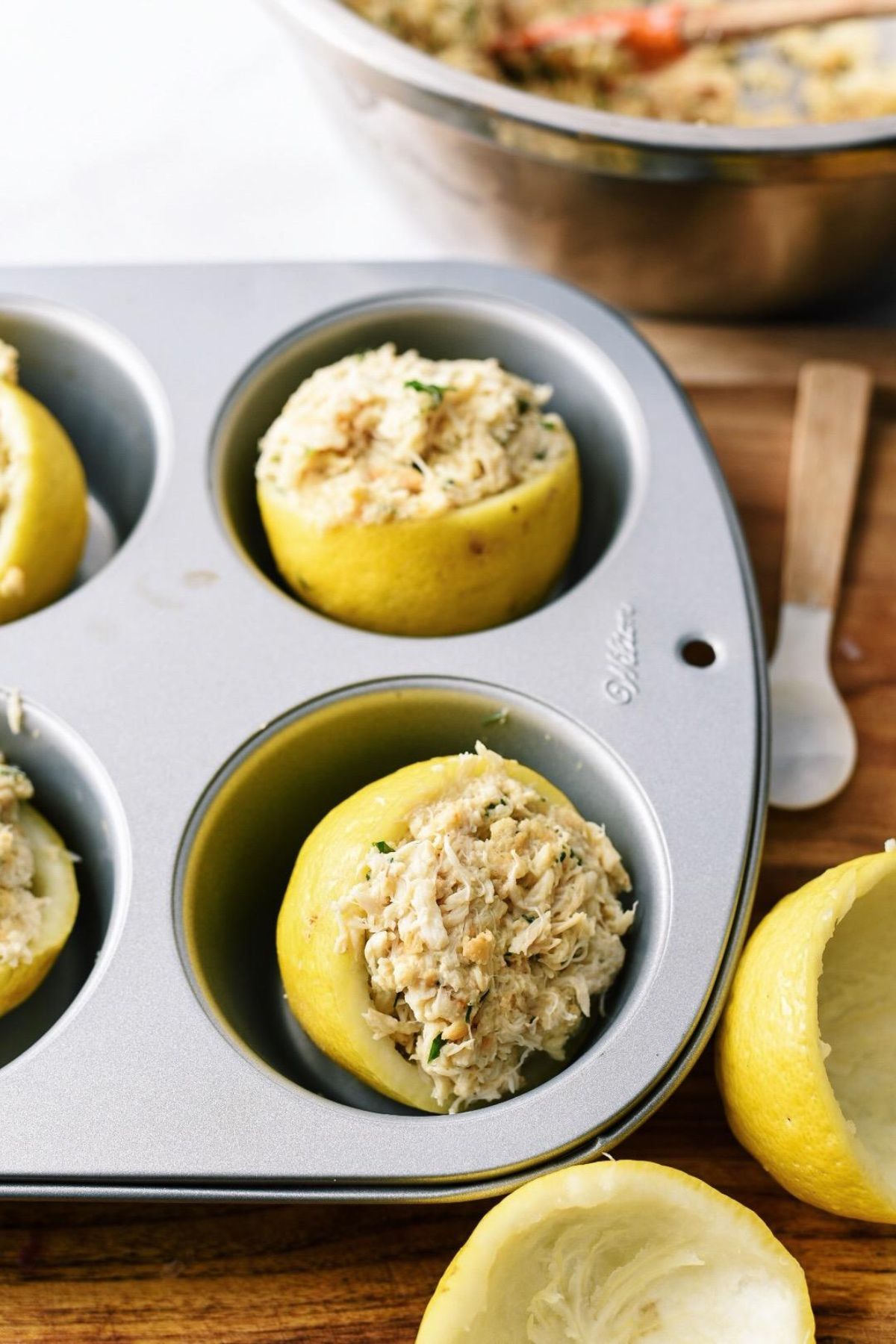 Crab cakes stuffed into lemon shells inside of a muffin tin, with empty lemon shells outside of the tin