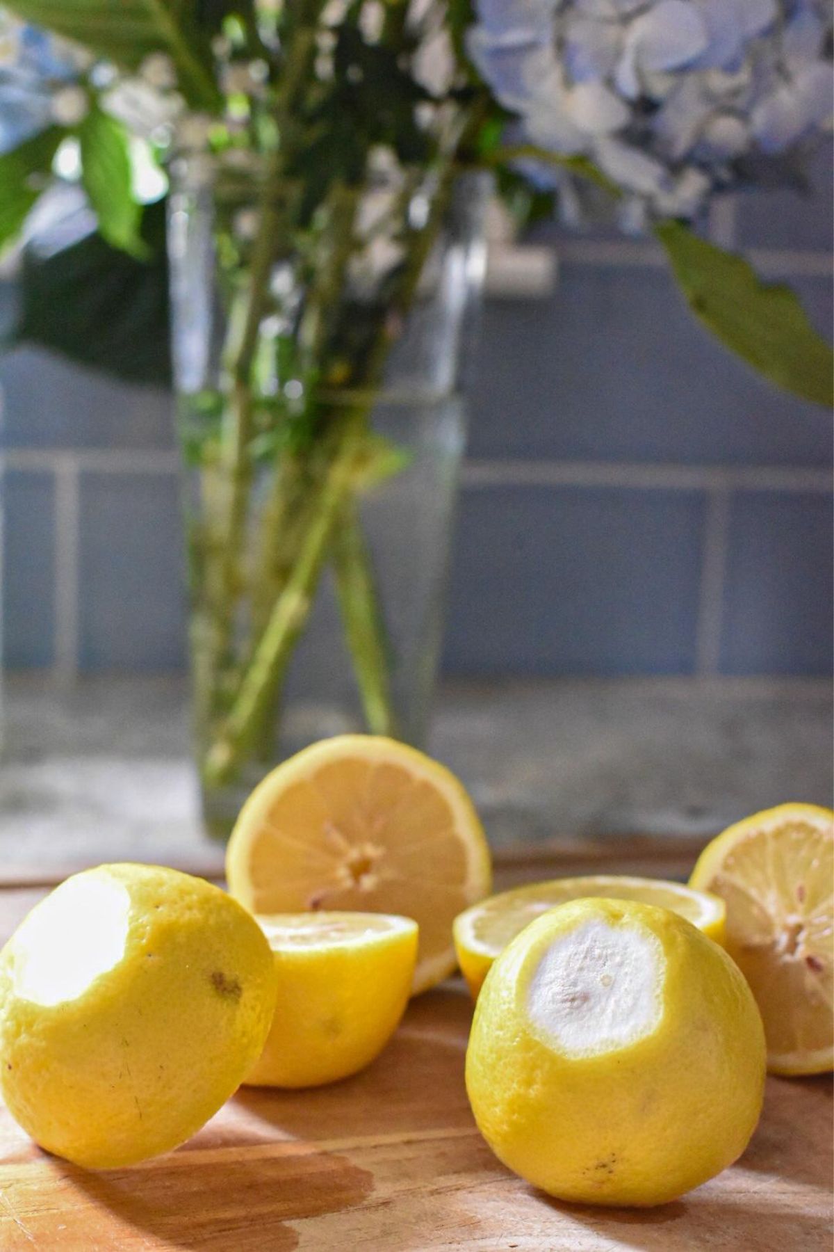 A bunch of lemons on a cutting board with the bottom sliced off, and a vase filled with folwers in the background in front of blue subway tile