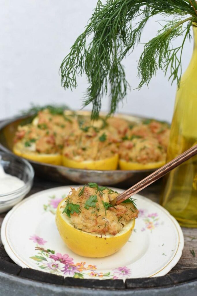 One stuffed lemon crab cake on a vintage tea cup plate, on top of an old whisky barrel, with other crab cake stuffed lemons in the background