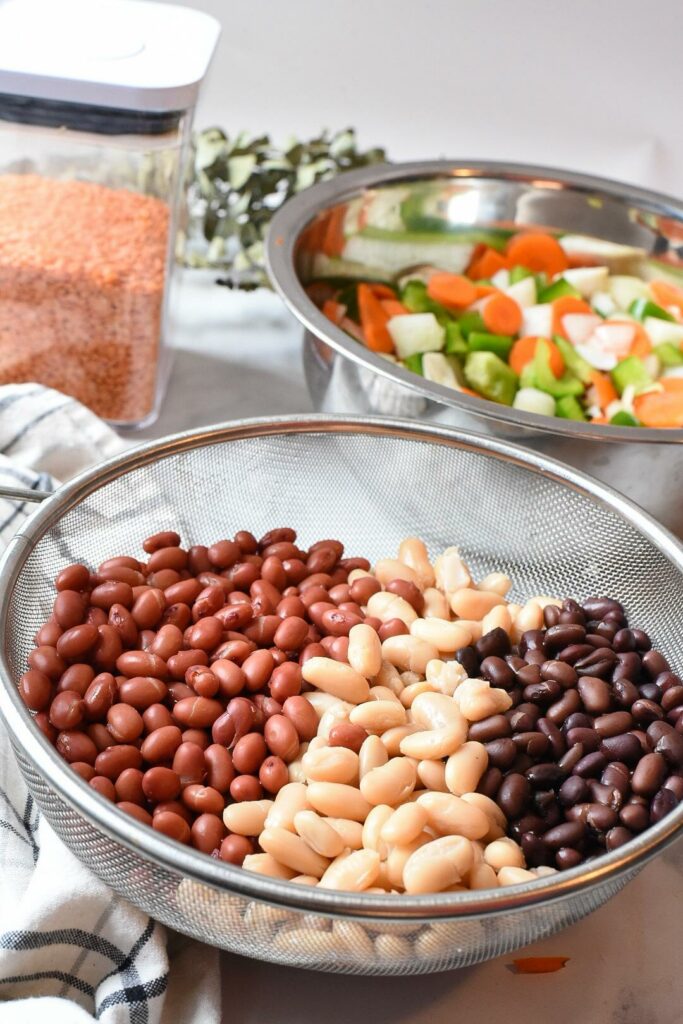 A mesh colander filled with three types of beans, and fresh chopped vegetables for vegan chili in the background.