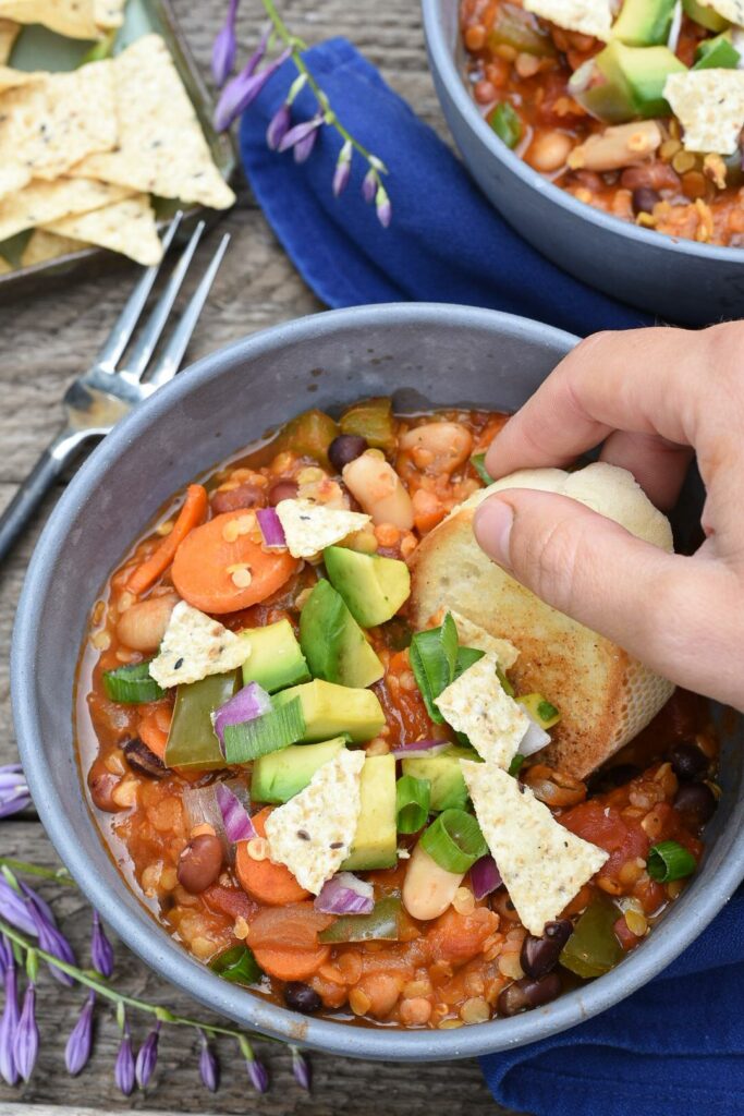 A hand dipping toasted bread into a grey bowl filled with the ultimate vegan chili recipe, topped with avocado and tortilla chips.