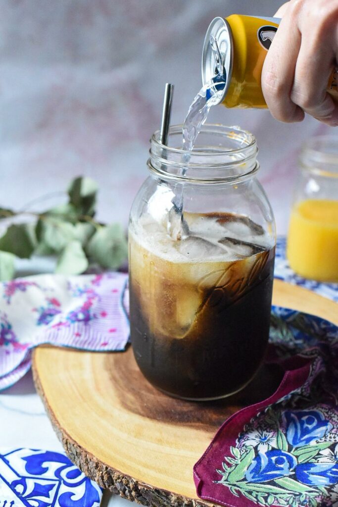 A large mason jar on wood with a hand pouring tonic water into espresso or coffee