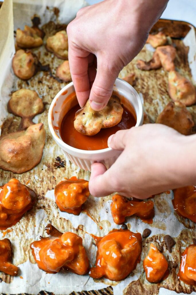 A hand dipping a frozen cauliflower floret into buffalo sauce, with a sheet pan filled with buffalo cauliflower wings under it
