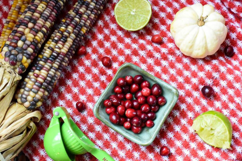 A red table cloth topped with cranberries, dried corn, limes and a white pumpkin