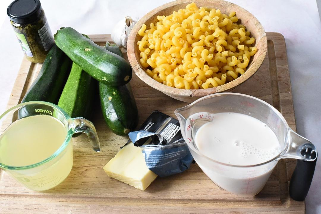 4 Zucchini, a bowl of pasta, milk, cheese, broth and pesto on a wooden cutting board.