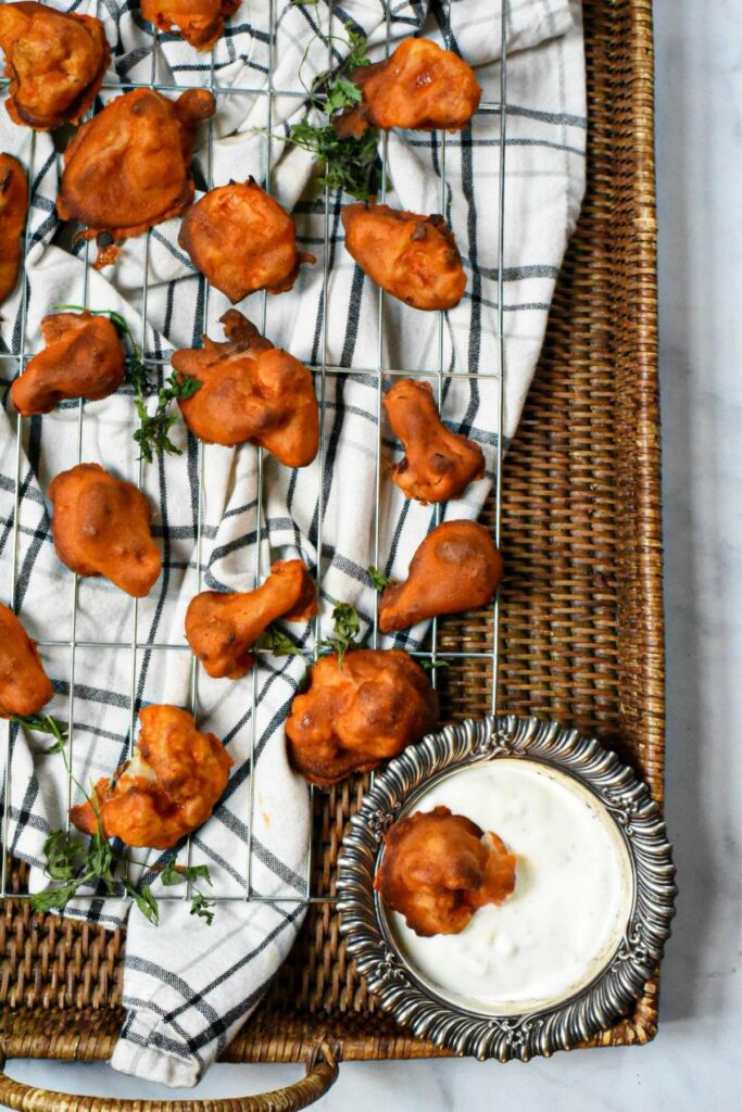 An overhead image of half of a metal baking rack filled with bright red buffalo cauliflower bites, with parsley sprinkled over the top and a wicker serving tray underneath 