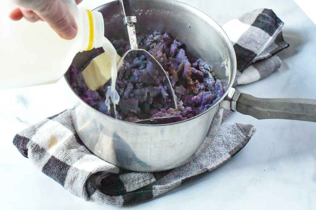 A pot filled with mashed purple potatoes, a potato masher and a block of butter, with a hand pouring milk into the pot.