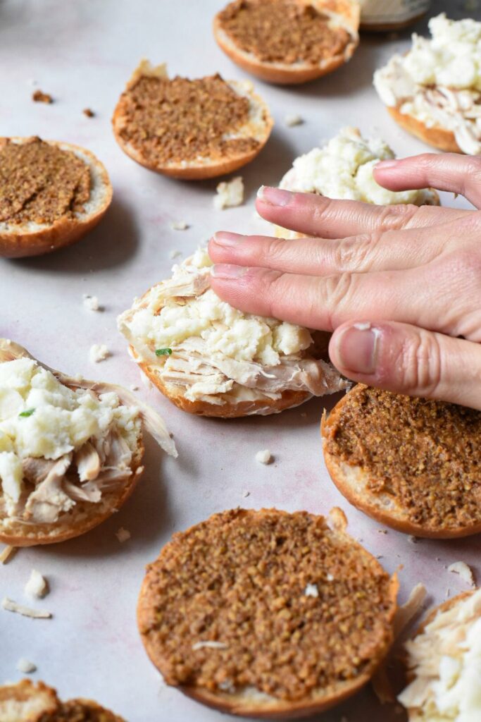 A hand patting mashed potatoes onto a turkey slider, with grainy mustard seen on the tops of the sliders.