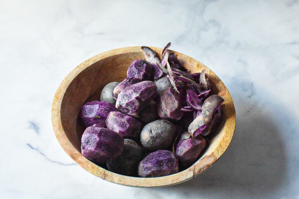 A wooden bowl on a white surface filled with bright purple potatoes, with some potatoes peeled and some not peeled.