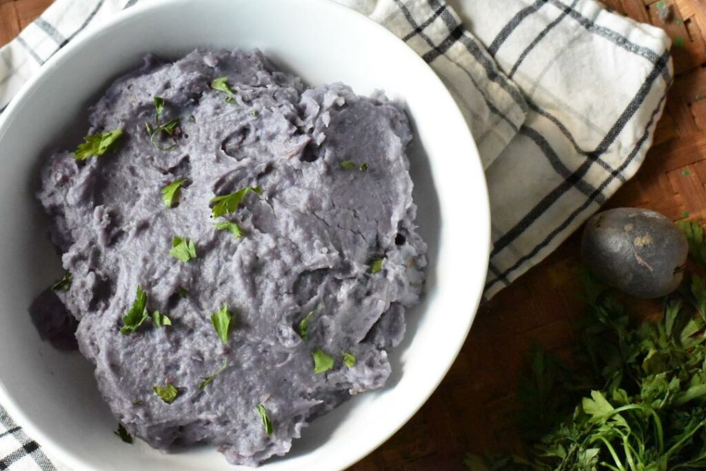 An overhead shot of a white bowl filled with purple mashed potatoes, sprinkled with parsley on top of a brown wicker plate, with a black and white plaid napkin under the bowl, and parsley in the corner.