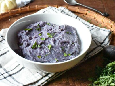 A white bowl filled with bright purple mashed potatoes on a wicker plate with a white plaid napkin under it.