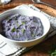 A white bowl filled with bright purple mashed potatoes on a wicker plate with a white plaid napkin under it.