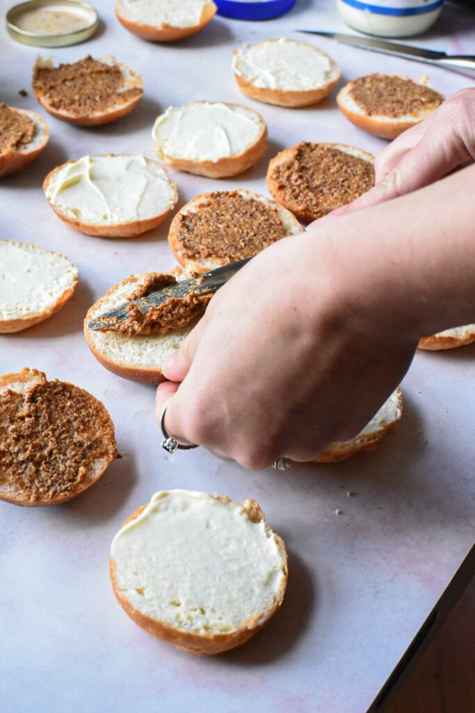 Hands spreading mustard on the tops of dinner rolls, with mayonnaise seen on the bottom of the dinner rolls.