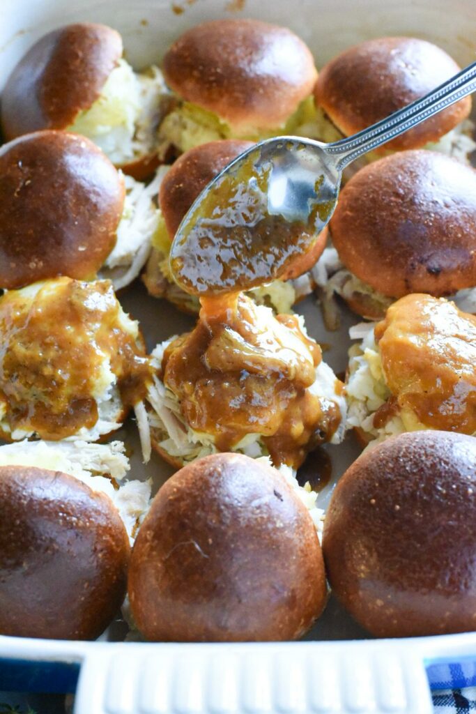 12 turkey sliders inside of baking dish, with a spoon pouring gravy over the top of three open faced turkey sandwiches.