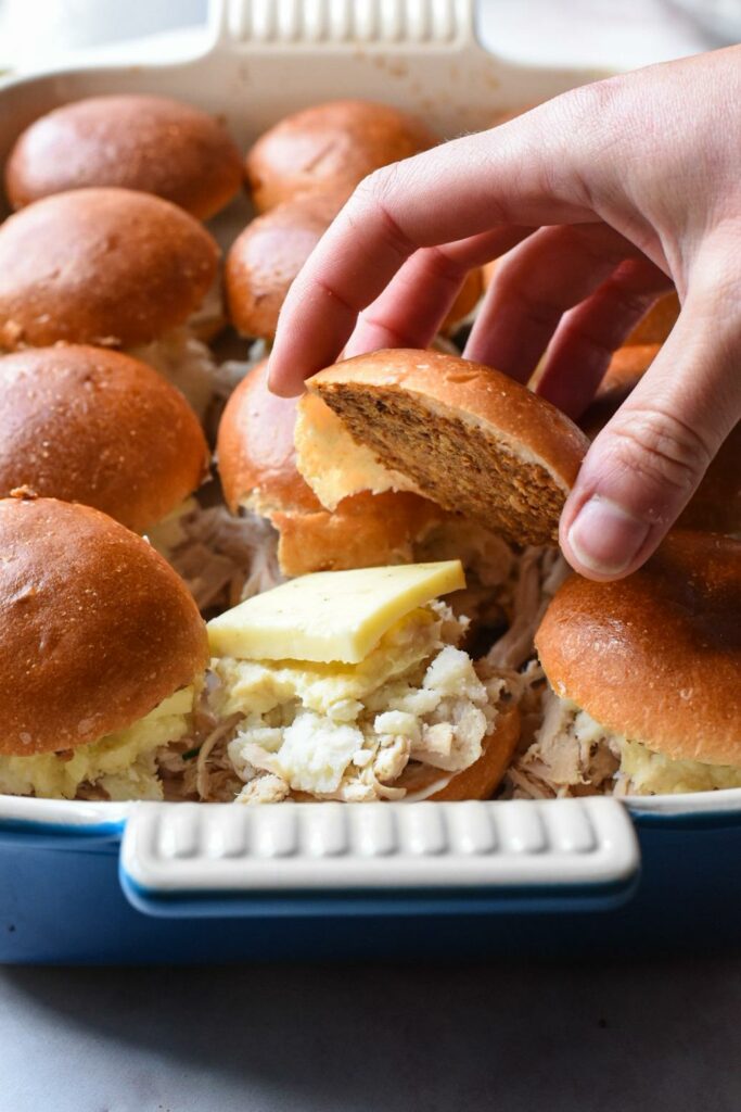A hand placing the top bun on a turkey slider inside of a baking dish, with a piece of cheddar cheese on top of the sandwich.
