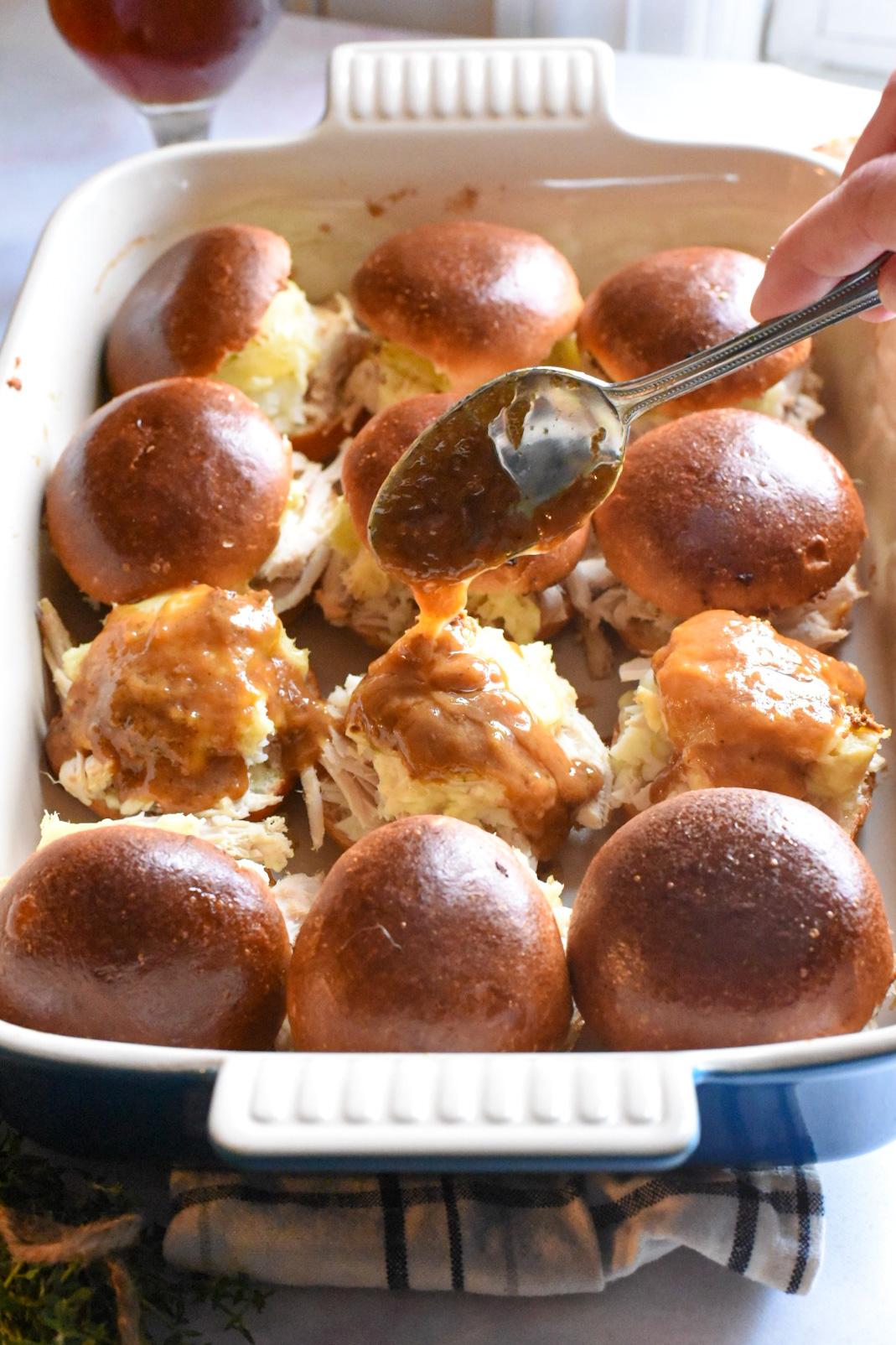 12 baked turkey sliders inside of a baking dish, with a spoon pouring gravy over three of the open faced sliders.