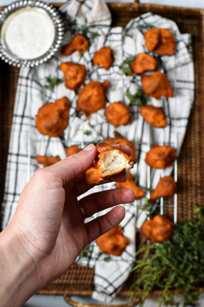 A hand holding a previously frozen buffalo cauliflower wing that has a bite taken out of it.  In the background there are more buffalo cauliflower bites on a wire baking tray with a black and white towel underneath.  