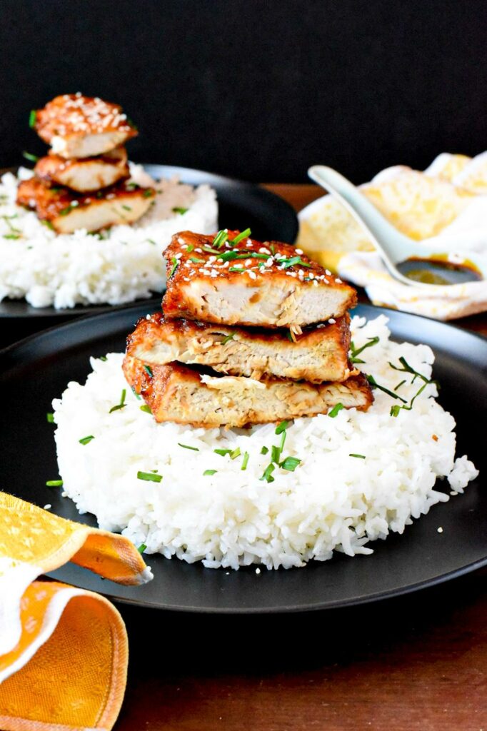 fried chicken breasts cut in half and stacked on top of white rice on a black plate.