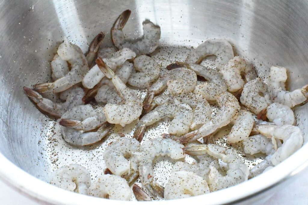 Raw shrimp in a metal bowl sprinkled with pepper and salt.