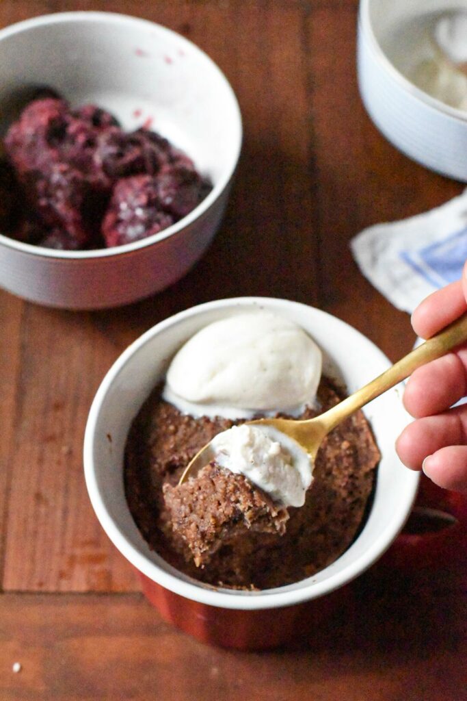 A mug filled with cherry mug cake, with a scoop of ice cream on top and a gold spoon inside the cake.
