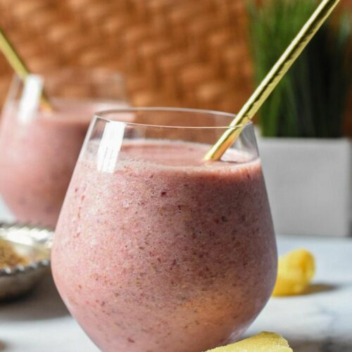 Close image of a glass filled with a pink smoothie with a gold paper straw coming out. There is another smoothie blurred in the background, and pieces of frozen pineapple on the white surface, with a wicker decoration behind the glasses.