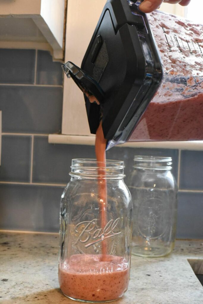 A pink anti inflammatory smoothie being poured out of a blender and into a large mason jar, with blue subway tiles in the background
