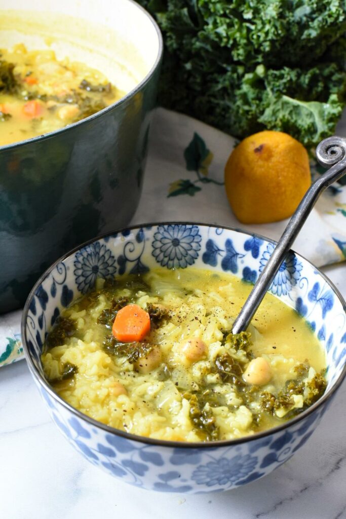 a blue and white flowered bowl filled with greek lemon soup with rice, kale, chickpeas and carrots visible