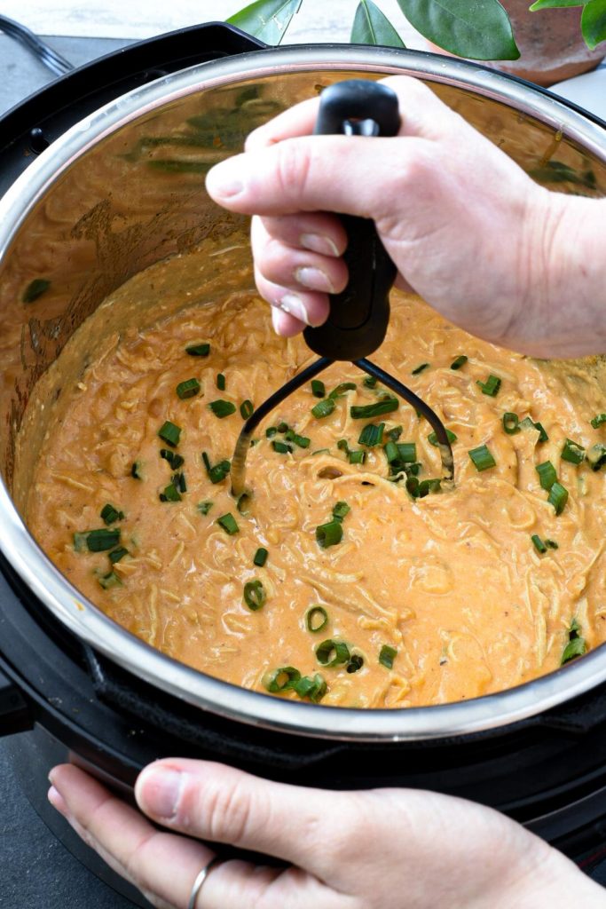 A hand holding a hand held masher mashing up buffalo chicken dip in an instant pot with green onion sprinkled over the top