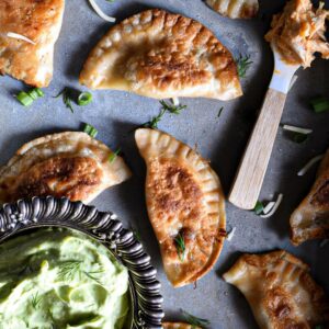 A bunch of buffalo chicken pierogies that have been pan seared until golden brown on a metal serving dish. There is a small spoon topped with buffalo chicken dip, and a metal bowl filled with avocado dipping sauce.