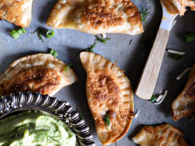 A bunch of buffalo chicken pierogies that have been pan seared until golden brown on a metal serving dish. There is a small spoon topped with buffalo chicken dip, and a metal bowl filled with avocado dipping sauce.