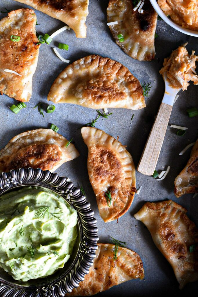 A bunch of buffalo chicken pierogies that have been pan seared until golden brown on a metal serving dish. There is a small spoon topped with buffalo chicken dip, and a metal bowl filled with avocado dipping sauce.