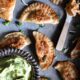 A bunch of buffalo chicken pierogies that have been pan seared until golden brown on a metal serving dish. There is a small spoon topped with buffalo chicken dip, and a metal bowl filled with avocado dipping sauce.
