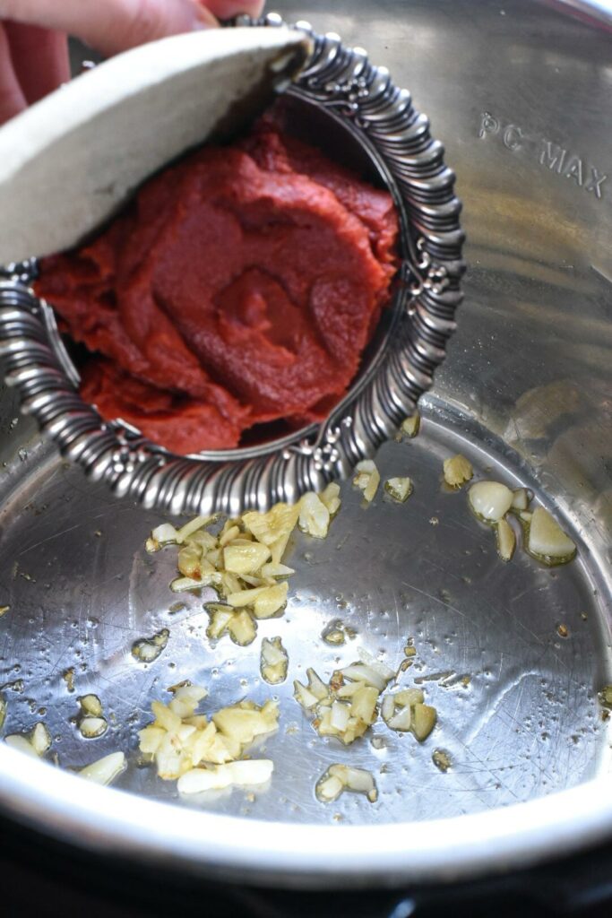 Tomato paste being poured into an instant pot that has garlic and ginger sauteing in it