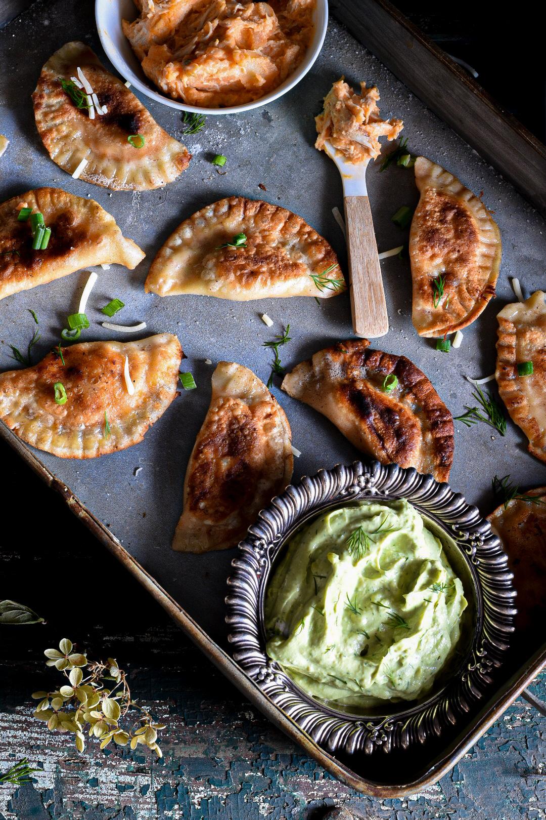 A close shot of a metal serving dish filled with golden crispy buffalo chicken pierogies with a metal bowl filled with avocado dip next to them and a spoon topped with buffalo chicken dip.