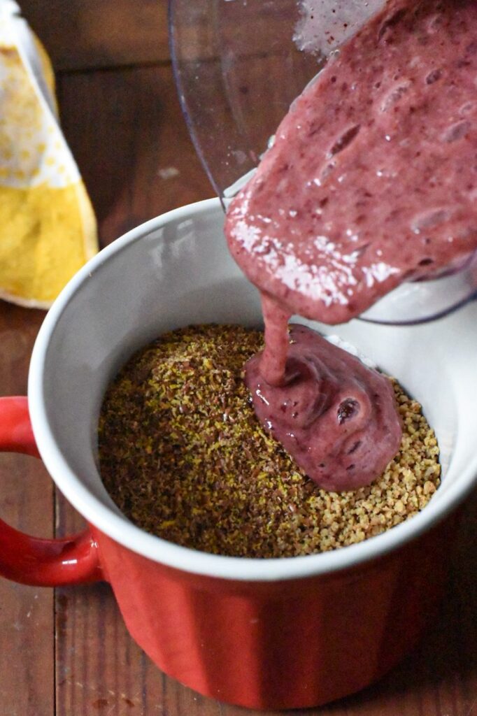 A blended pink cherry mixture being poured into a mug filled with flaxseed meal. 