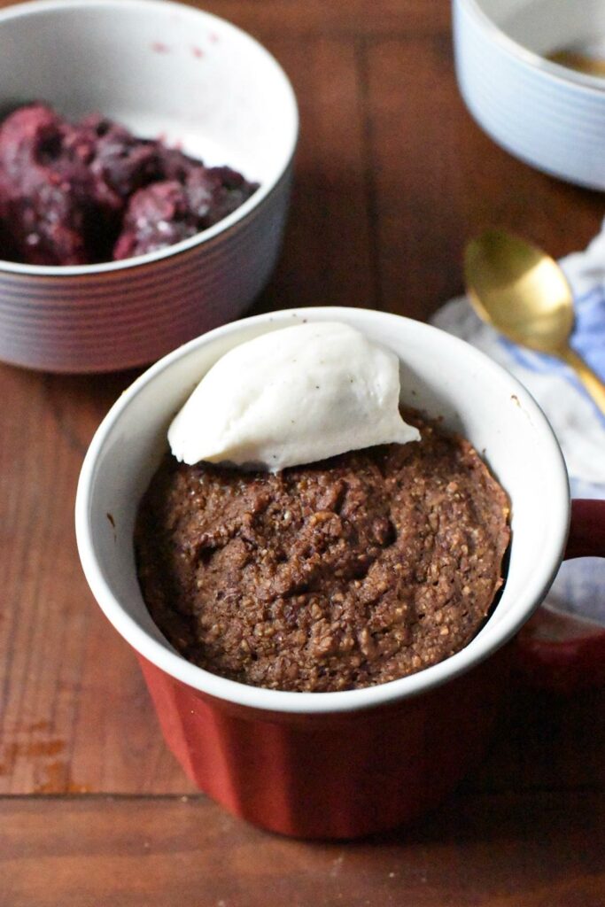 A sugar free mug cake in a red mug with a dollop of vanilla ice cream on top, and a gold spoon in the background.