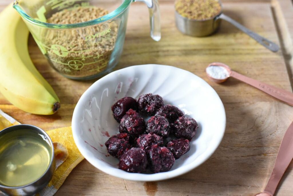 A wooden cutting board with a bowl of frozen tart cherries, a banana, a measuring cup filled with flaxseed meal and a gold measuring spoon filled with baking powder.