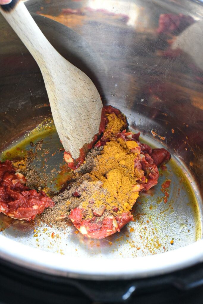 Spices and tomato paste being stirring by a wooden spoon inside of an instant pot