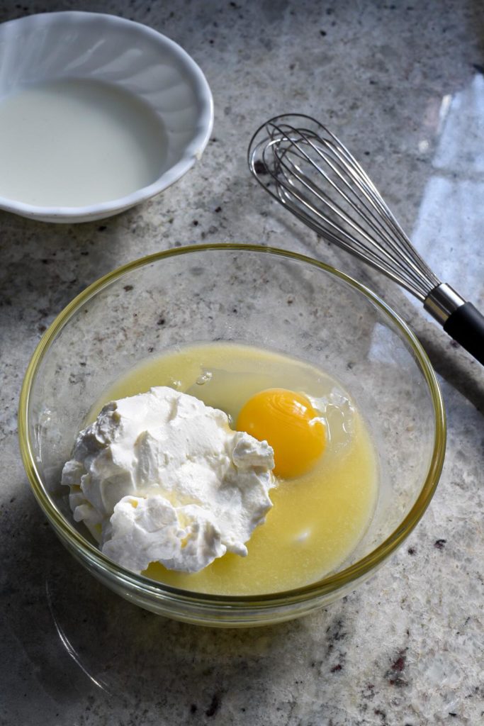 A glass bowl filled with a raw egg and greek yogurt, with a whisk behind it.