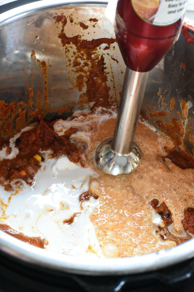 A hand held blender inside of an instant pot blending cream and tomato sauce