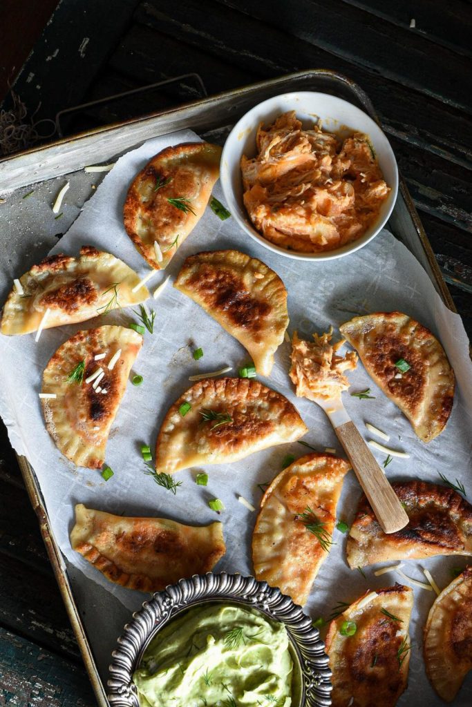 An overhead shot of a metal serving dish filled with golden crispy buffalo chicken pierogies with a metal bowl filled with avocado dip next to them and a spoon topped with buffalo chicken dip.