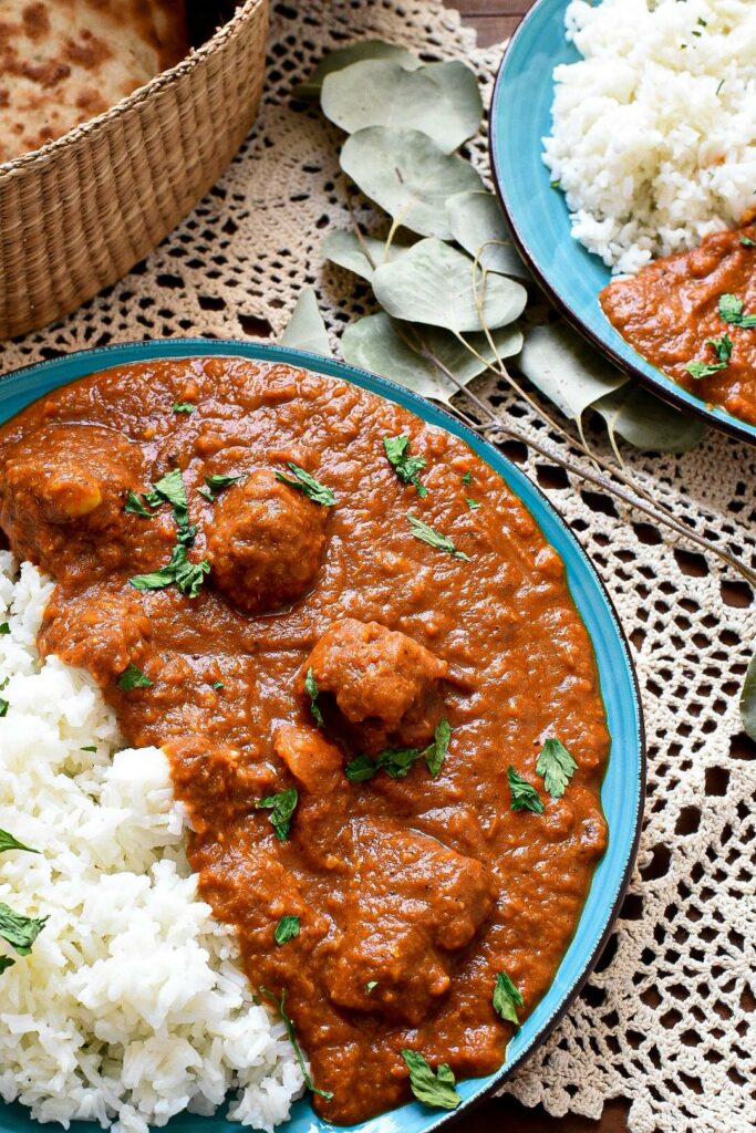 Two blue plates on lace filled with a bright red cauliflower tikka masala sauce, with basmati rice on the plates and naan in the background in a wicker basket