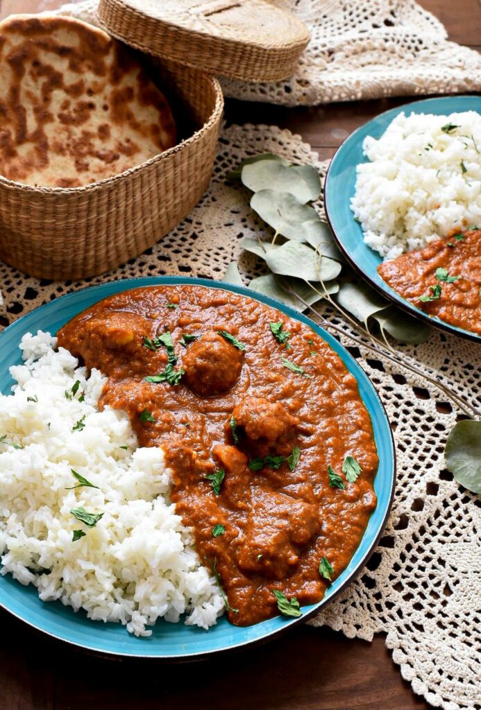Two blue plates on lace filled with a bright red cauliflower tikka masala sauce, with basmati rice on the plates and naan in the background in a wicker basket