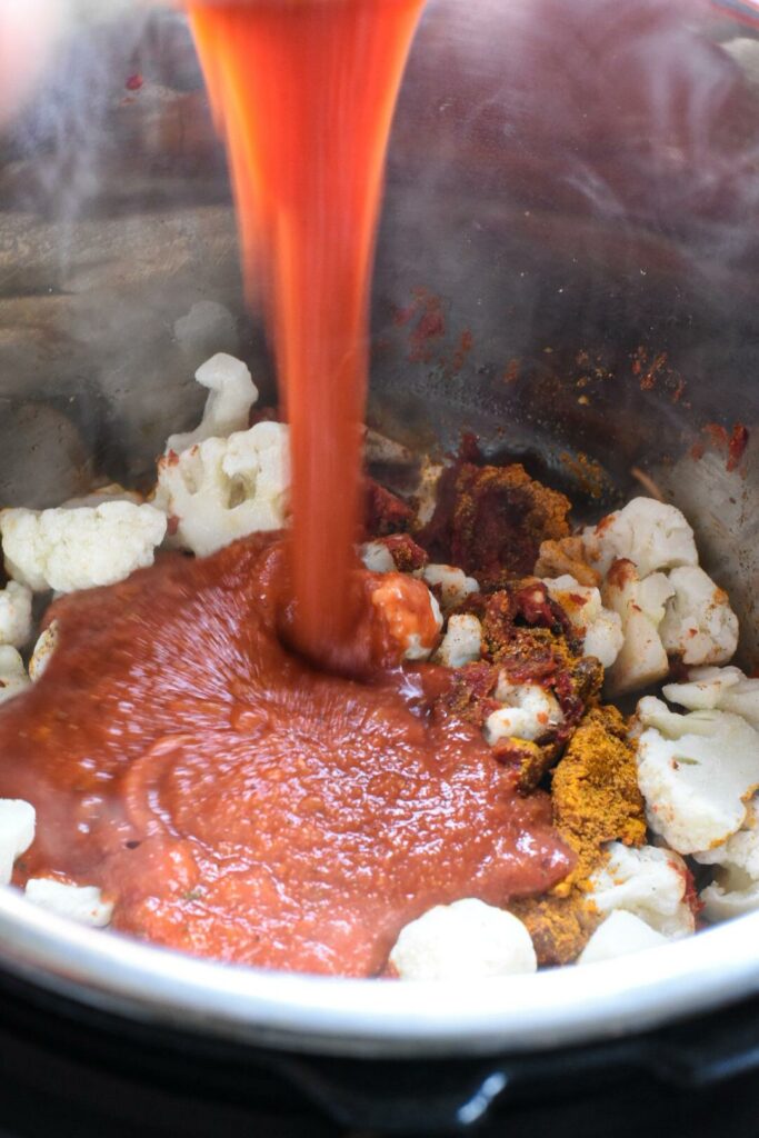 Tomato sauce being poured into an instant pot filled with cauliflower, spices and tomato paste